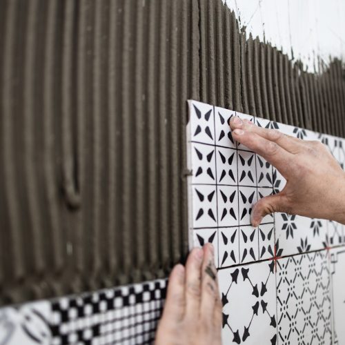 A technician carefully places beautiful patterned tiles on the wall in a home.